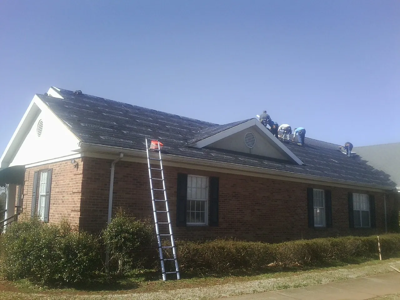 Crew tearing shingles off a brick ranch roof with a ladder set — Emergency Roof Repair work by our crew in Mulliken