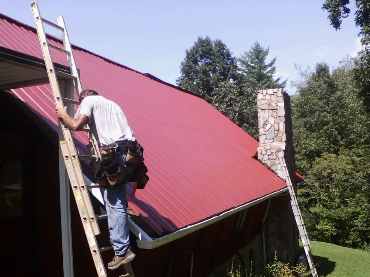 Roofer on a ladder beside a red metal roof and stone chimney — Soffit & Fascia Repair by Roofing Brothers in Mulliken